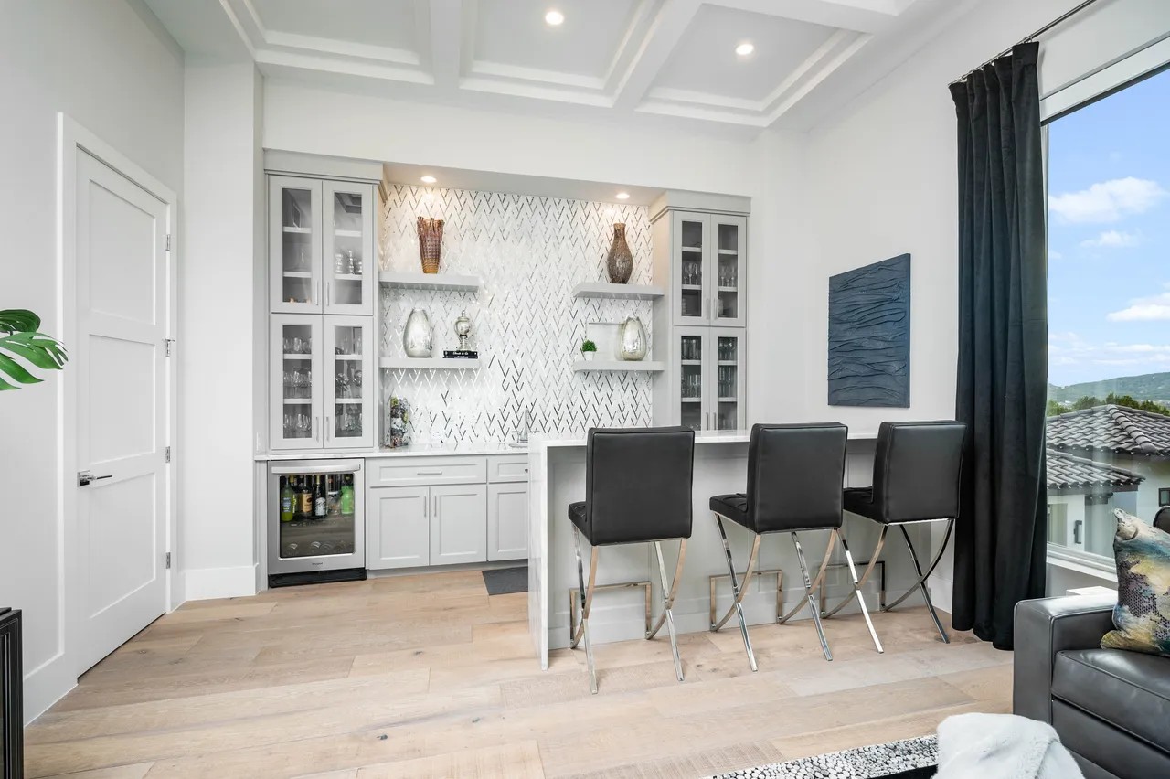 Custom home bar with white painted cabinetry, herringbone marble backsplash, floating shelves, and glass-front upper cabinets by Amaya