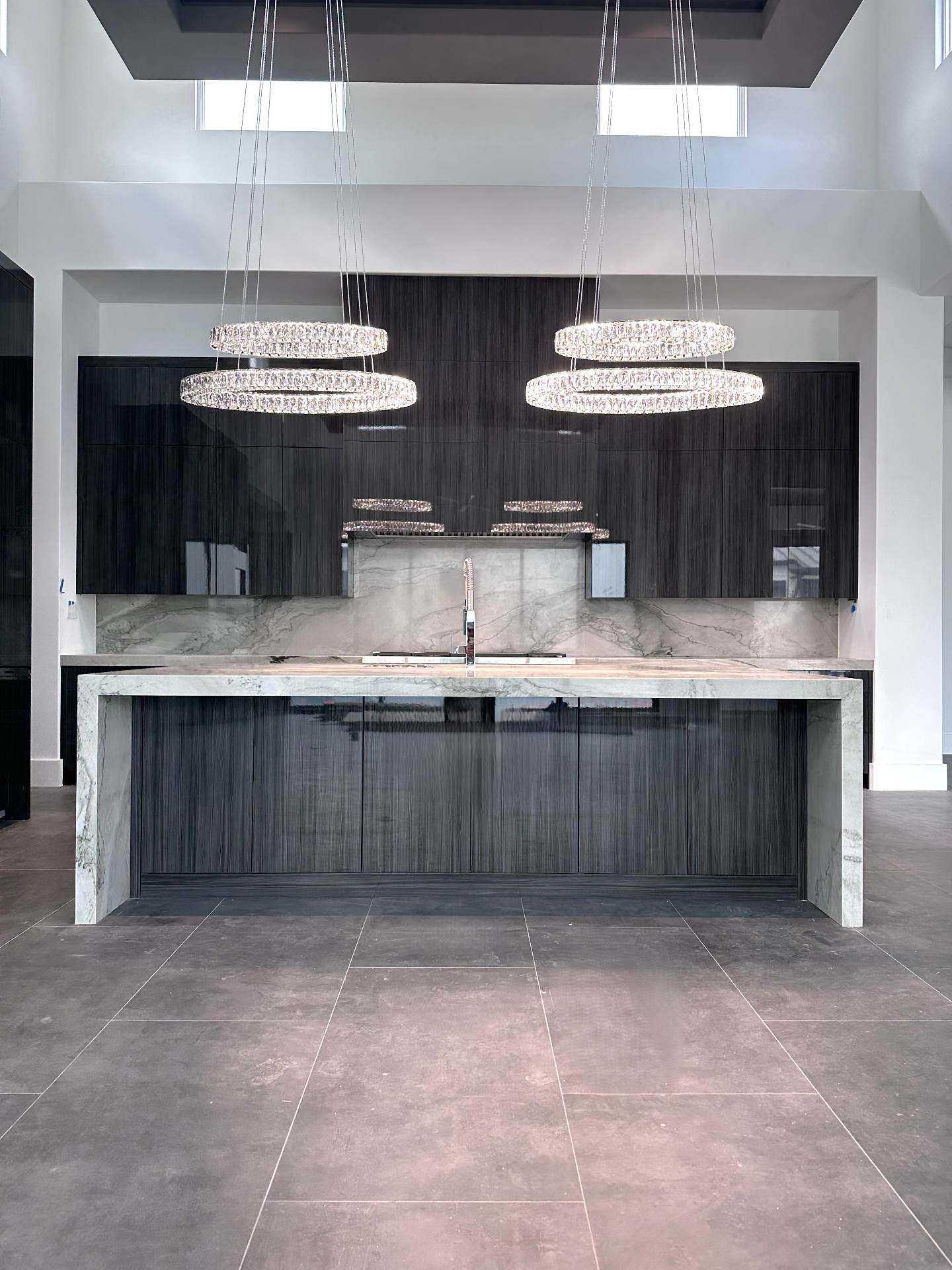 Modern high-gloss kitchen with linear dark cabinetry, quartzite waterfall island, and layered crystal ring chandeliers by Amaya
