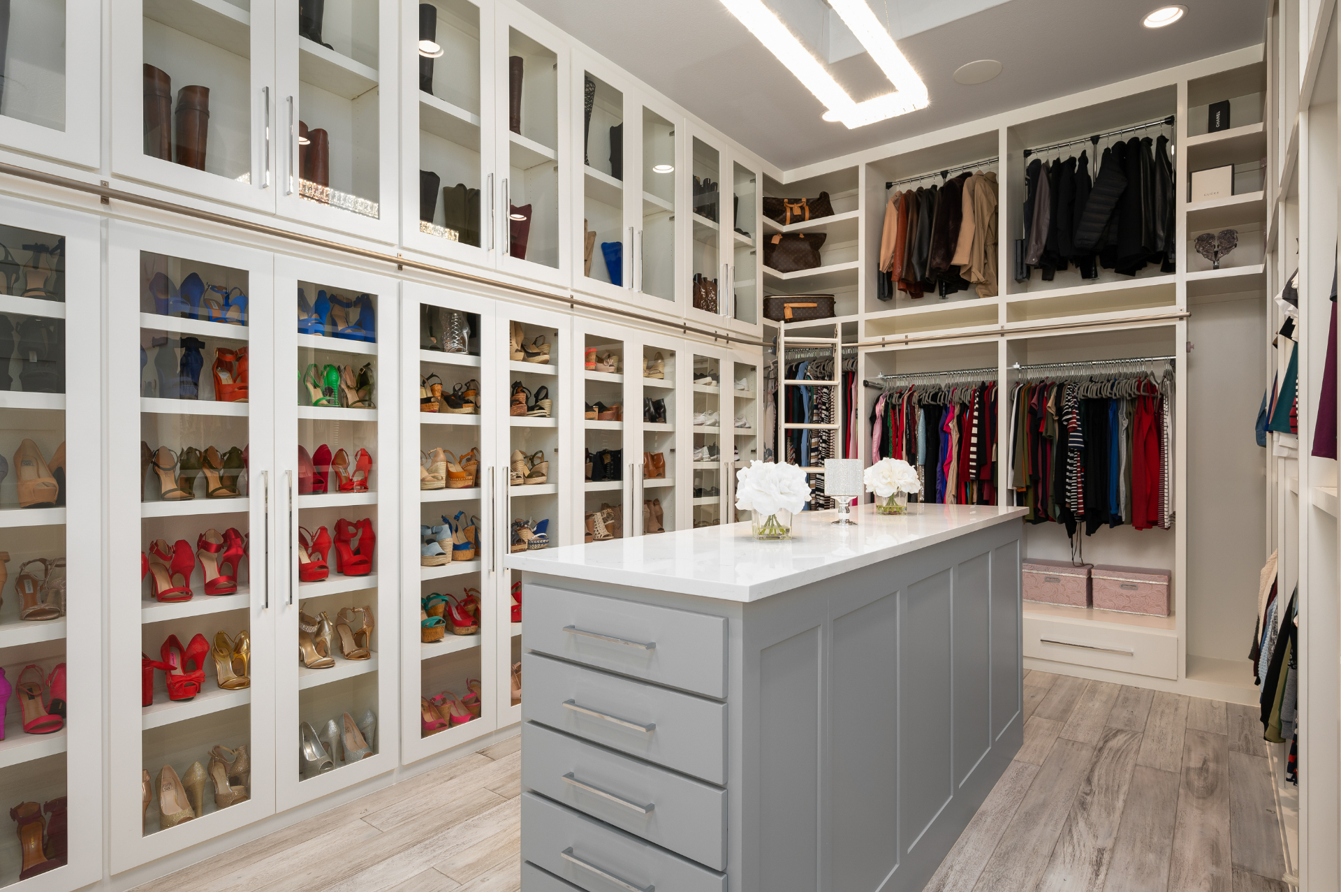Bright white walk-in closet with glass-front cabinetry, illuminated shoe display, and a slate blue island by Amaya