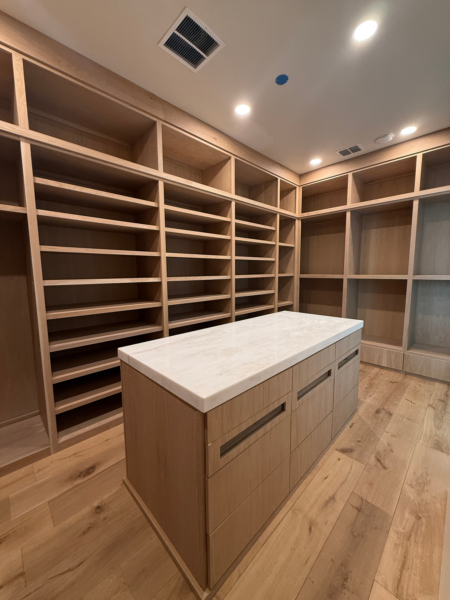 Custom walk-in closet with floor-to-ceiling white oak shelving and a marble-topped island dresser by Amaya